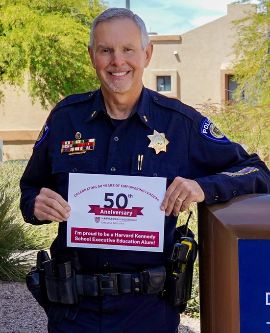 Bob Sommerfeld in a police uniform, holding a sign celebrating the 50th Anniversary at Harvard Kennedy School Executive Education, reading, "I'm proud to be a Harvard Kennedy School Executive Education Alum!"