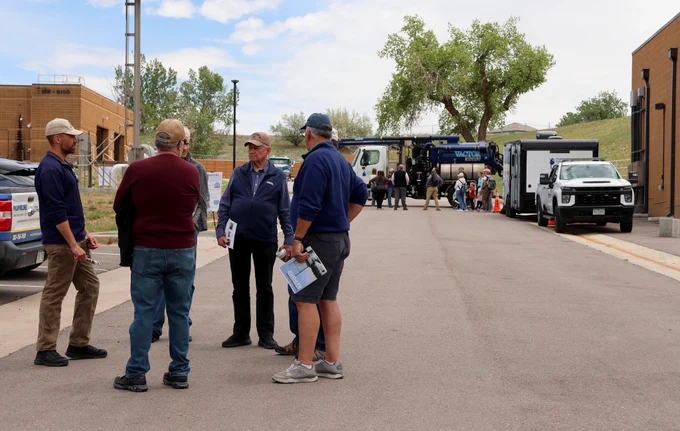 Group of six men standing in a circle talking during a tour at a wastewater treatment plant with another group in the background standing near a vacuum truck.