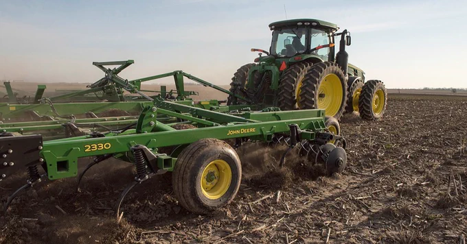 A John Deere tractor is shown working in a field, pulling a large piece of farming equipment with the number 2330 printed on it, turning the soil. The sky is clear, and the field is mostly bare.