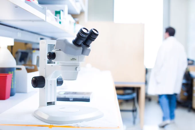 Close-up of a binocular microscope on a lab bench with a scientist in a white coat blurred in the background.