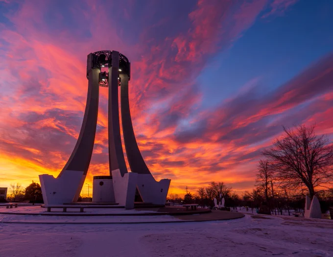 A dramatic sunset casts vibrant hues of orange, pink, and purple across the sky, silhouetting the United Nations Monument in Rapid City, South Dakota. Snow covers the ground, and bare trees are visible on either side of the monument.