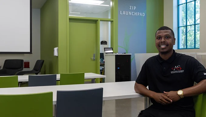 A person is sitting on a chair in a modern classroom with green chairs and white tables. The background features a sign that reads "ZIP Launchpad." The room is bright with large windows to the right.