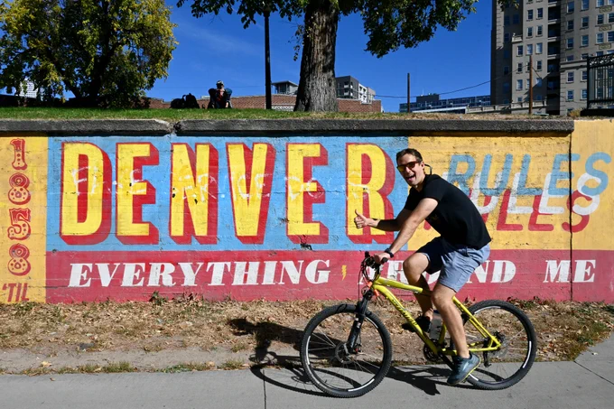 A man in sunglasses, a shirt and shorts bikes down the Cherry Creek Trail on a sunny day. He gives a thumbs up and a big smile as he rides past a colorful mural that reads in all caps "Denver rules everything around me".
