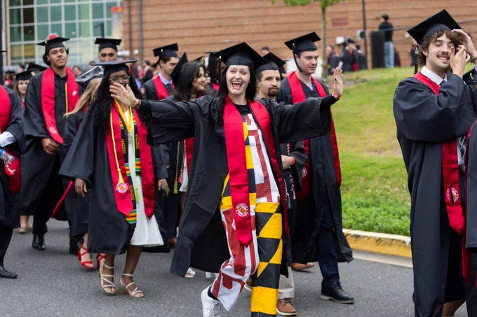 A student wearing graduation regalia and Maryland flag overalls poses enthusiastically amidst a processional of graduating students.