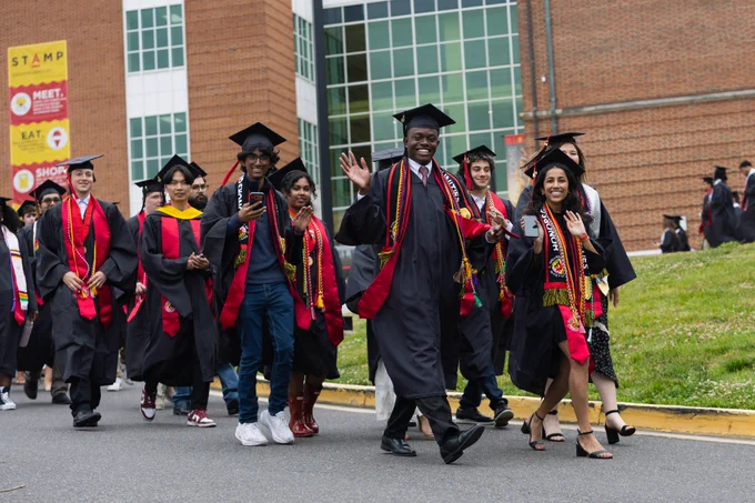 Students wearing graduation regalia wave and smile at the camera during a processional of graduating students.