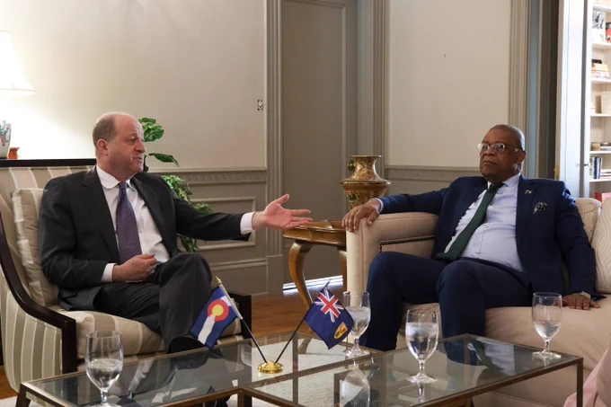 Two men in suits engaged in a formal discussion, seated in a well-lit room with small flags of Colorado and Turks and Caicos on the glass table between them.