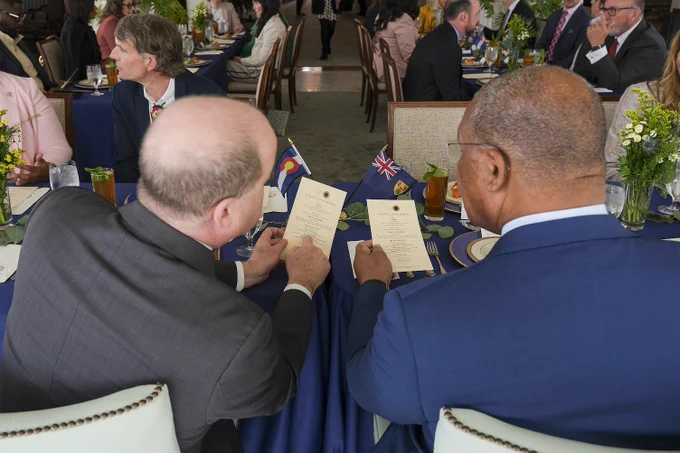 Back view of two men examining papers at a blue-clothed table during a formal event with guests and floral centerpieces in the background.
