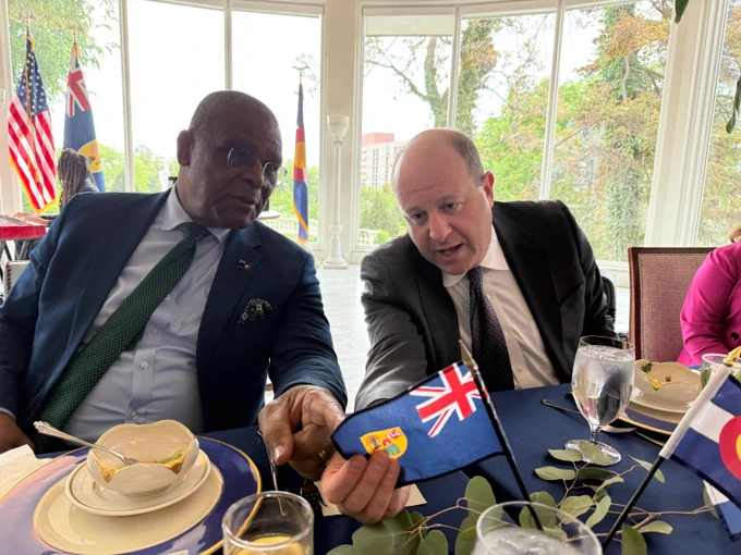 At a formal event, two men in suits sit by a windowed room, one displaying a small flag of Turks and Caicos