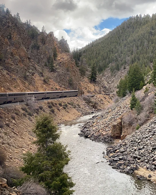 Amtrak train winding through a rocky mountain canyon beside a river, surrounded by pine trees under cloudy skies.