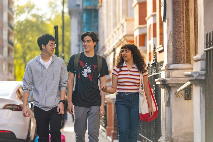 Three students stroll down a London sidewalk, engaged in conversation. The woman, with curly hair and a striped shirt, gestures as she speaks. Buildings and a parked car line the street.