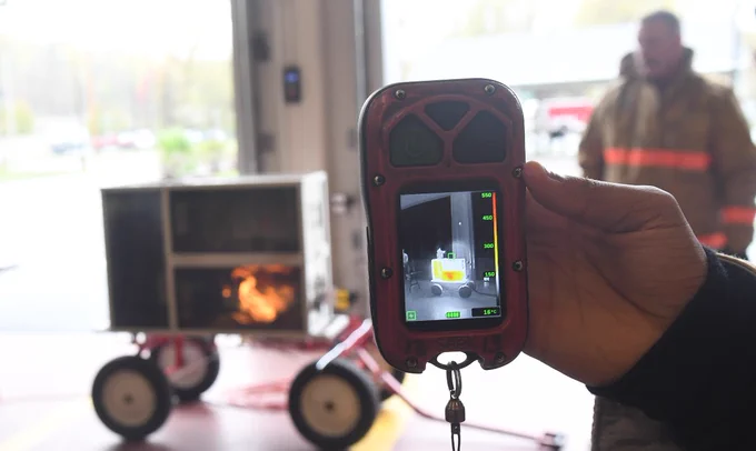 Hand holding a thermal imaging camera showing a heat signature of a fire inside a metal box on wheels in a garage.