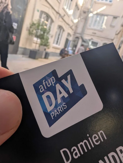 A hand holds a badge featuring "AFUP Day Paris" in front of a busy street with people and buildings in the background.