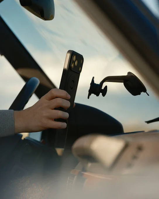 A hand holds a smartphone with visible camera lenses and attachments, in front of a blurred background.