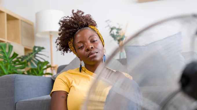 Woman in a yellow top sat in front of a fan that is blowing cool air in her face.
