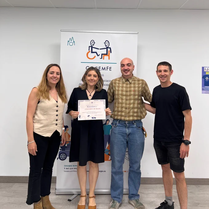 Foto de familia de los equipos técnicos de Integra CEE y Cocemfe Burgos. Una de las profesionales de la entidad social, en el centro de la imagen, posa con un diploma de reconocimiento. Detrás de los cuatro profesionales, un roll up con la imagen corporativa de COCEMFE Burgos.