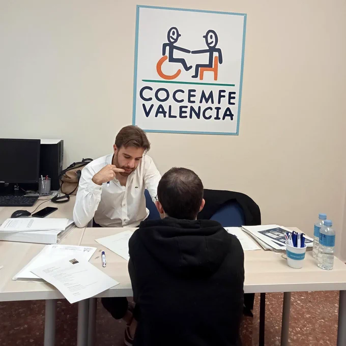 Momento de una entrevista presencial en las oficinas de Cocemfe Valencia. Un jefe de servicio de Integra CEE charla con un usuario de la entidad social. Los dos están sentados de frente en una mesa rectangular de oficina. Al fondo, sobre la pared, un cartel con el logo de Cocemfe Valencia. 