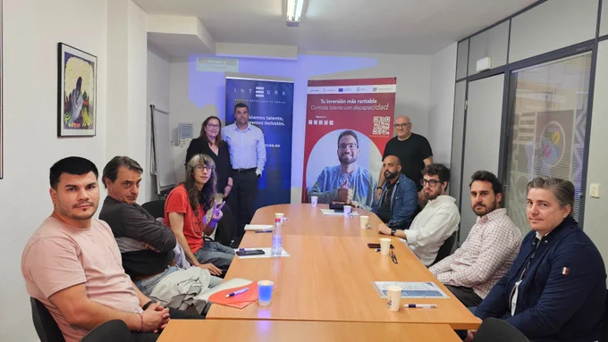 Sala de reuniones con diez personas sentadas alrededor de una mesa. Al fondo se ven roll ups de Integra CEE e Inserta Empleo con el mensaje sobre contratación de talento con discapacidad. La mesa tiene documentos, bolígrafos y vasos.