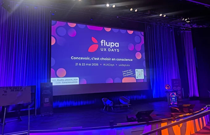 A stage set for UX Days with a colorful backdrop, featuring event details, a podium, and two chairs. The atmosphere is vibrant and professional.