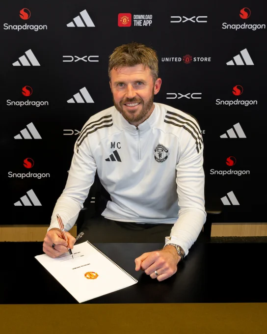 Michael Carrick in a white training top signing a contract at a table with branded backdrop behind him.