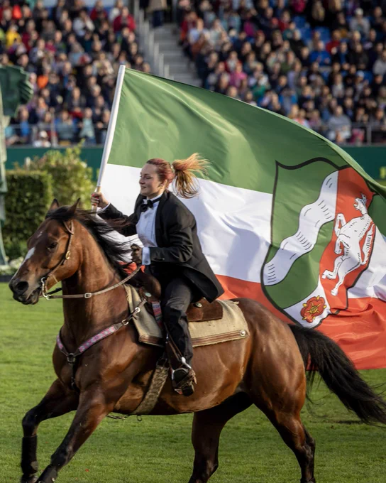 Eine Frau in einem schwarzen Anzug reitet auf einem braunen Pferd und hält in ihrer linken Hand eine Nordrhein-Westfalen Flagge.