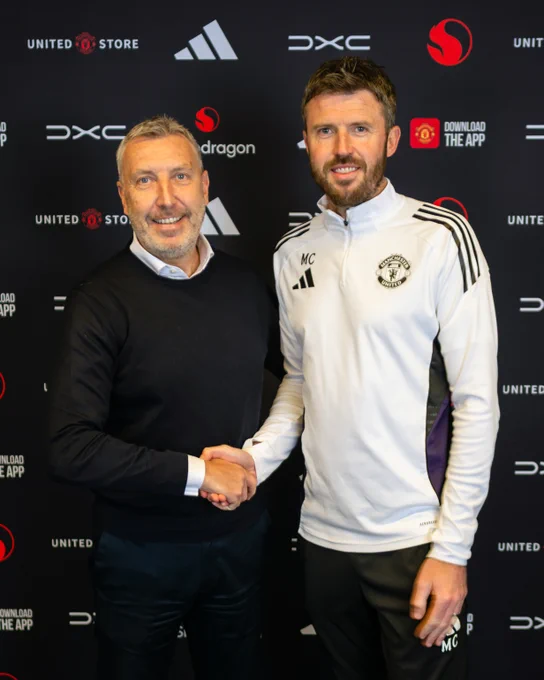 Jason Wilcox and Michael Carrick in front of a backdrop featuring Manchester United and sponsor logos, Wilcox wearing a black jumper and Carrick wearing a white Manchester United training top.