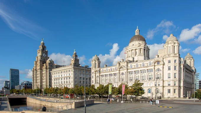 A wide daytime view of Liverpool’s waterfront featuring the Royal Liver Building with its clock tower on the left and the domed Port of Liverpool Building on the right. The historic white stone buildings line a tree-filled plaza with paved walkways, a few pedestrians, and flags on poles. The sky is bright blue with scattered clouds, and modern buildings are visible in the distance.