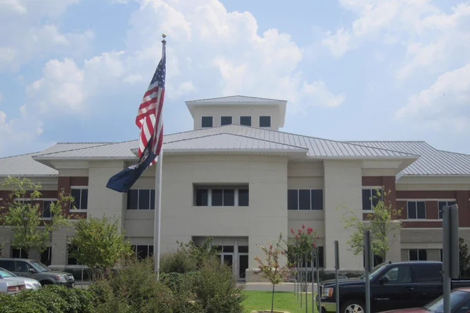 A modern, two-story building with a metal roof, an American flag and another flag in front, cars parked, and a partly cloudy sky overhead.