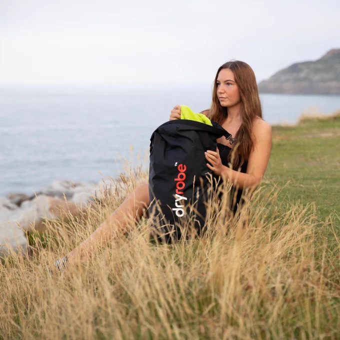 A woman sits on a grassy hill overlooking the ocean on an overcast day, packing a bright lime green towel into a black, waterproof Dryrobe compression bag. Dry grass is in the foreground, with coastal cliffs and calm grey sea stretching into the distance.