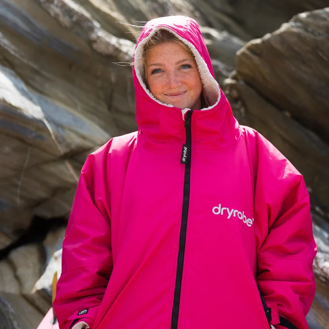 A close-up portrait of a smiling woman with freckles wearing a vibrant, hot pink Dryrobe Advance long-sleeve change robe. The hood is up, revealing a fluffy white fleece lining. The white dryrobe logo is printed on the left chest, and a rugged, grey rocky cliff face serves as the softly blurred background.
