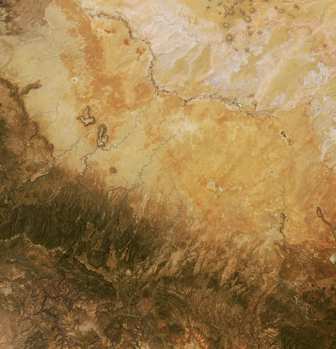 Landsat 8 image of the Arizona desert. The landscape is a flat brownish yellow in the upper right two thirds. Patches of rusty orange dot the surface. The lower left third turns rocky, with darker brown and green ridges running toward the corner. Thin ribbons of river run out from the rockier hills in several places. One larger river runs from about 12 to 3 o’clock, with patches of lighter white to the northeast of it.