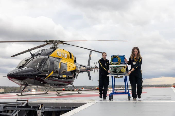 Two medical personnel in uniforms are transporting medical equipment on a wheeled cart across a helipad. A black and yellow helicopter is parked nearby under a cloudy sky, marked with "AirCare" and "Iowa." The scene suggests a medical emergency or transport situation.