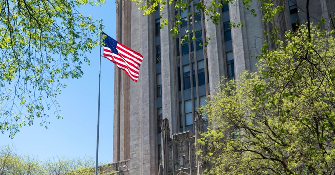 A tall American flag flutters next to a historic stone building, partially obscured by green trees. The sky is clear and blue.