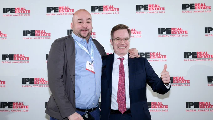 Two people posing together, smiling, one giving a thumbs-up. They stand in front of a IPE Real Estate Global Awards 2026 backdrop.