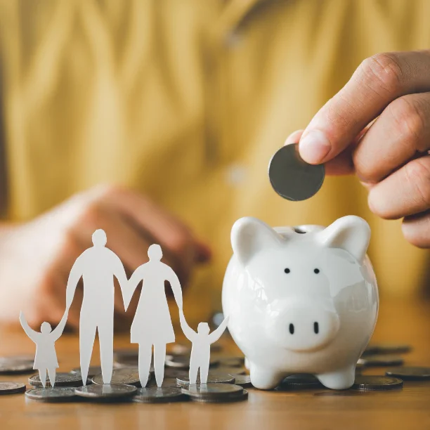 Hand placing a coin into a white piggy bank next to paper cutouts of a family on a wooden table with scattered coins.
