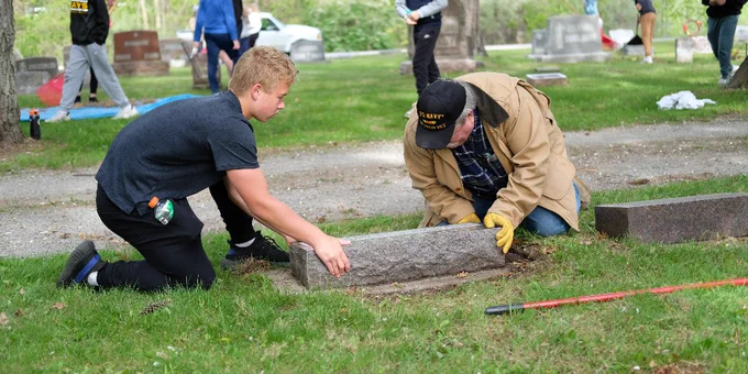 Two people carefully reposition a gravestone in a cemetery surrounded by trees and other headstones.