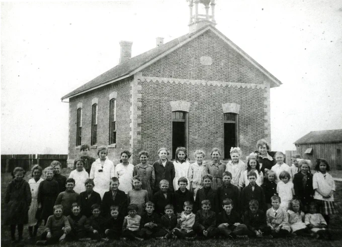 Black and white photo of a large group of children and adults posing in front of a brick schoolhouse with a bell tower.