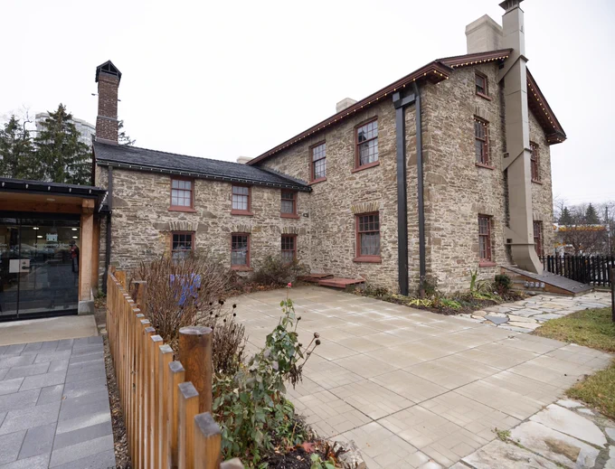 Historic stone building with red-trimmed windows and a chimney, surrounded by a small garden and wooden fence.