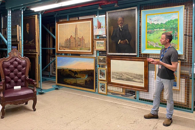Man standing in an art storage room, gesturing toward a collection of framed paintings and portraits on a metal grid wall.