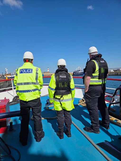 Three police officers in high-visibility jackets and hard hats on a boat deck with a harbour in the background.