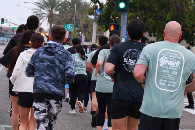 A group of runners supporting Special Olympics Southern California jogs together down a city street lined with palm trees, wearing matching event T-shirts as they pass through an intersection with a green light.
