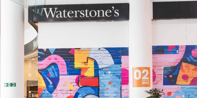 A colorful mural with abstract shapes and patterns is painted on a wall underneath a Waterstone's bookstore sign. An orange sign with "02 centre NW3" is visible on the right side of the image.