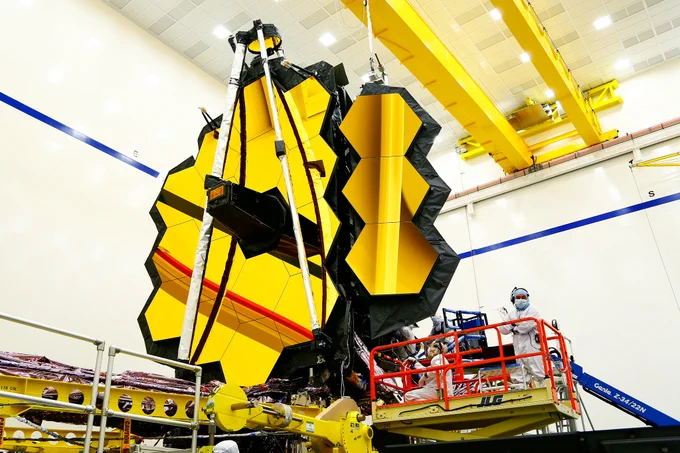 The Webb telescope in a clean room. Its golden primary mirror dominate the center of the image. Fifteen of the 18 hexagon mirror segments are deployed, with the 3 on the right folding into place. Together, they make a jagged hexagon shape. The secondary mirror support is folded up in front of the primary mirror so it points to the ceiling. Workers in head to toe white clean room suits are on a red left next to the mirrors.
