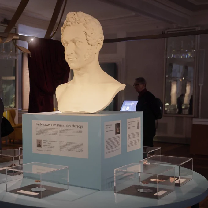 Installation aus der Ausstellung "Coburg | Gotha | 1826". Eine Büste steht auf einem Podest mit um sie gruppierten Objekten in Minivitrinen und Informationstafeln. Foto: Sergej Tan
