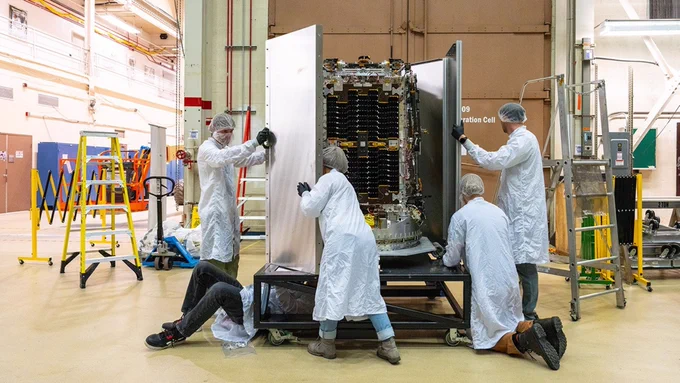 Four engineers in clean room suits stand and kneel around a refrigerator-sized metal box. They are in the process of opening the box, to reveal the LINK satellite. The visible part of the spacecraft is covered with the black grid of a solar panel. They are working on a large, open warehouse-type floor.
