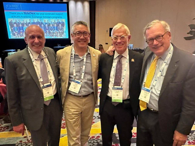 Four men in business attire, wearing nametags on lanyards, smile and pose for a photo
