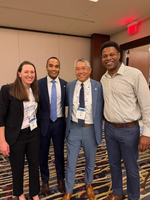 Four people in business attire, wearing nametags on lanyards, smile and pose for a photo
