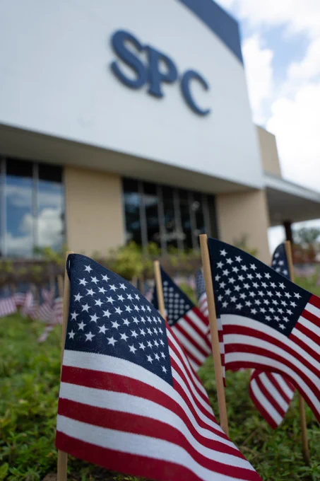 Small American flags are planted in the grass in front of a building with large letters reading "SPC". The sky is partly cloudy.