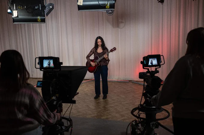 A person stands in a studio holding a guitar, surrounded by three camera operators recording the performance. The setting includes large wall curtains and studio lights overhead.