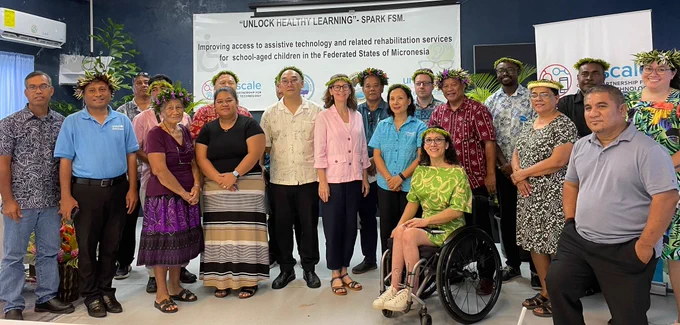 A group of people stand together indoors at the launch of the “Unlock Healthy Learning” SPARK FSM initiative in the Federated States of Micronesia. The group includes representatives from ATscale, UNICEF, government and community partners, many wearing traditional floral head garlands. A woman seated in a wheelchair is positioned at the front of the group. Behind them, a large banner reads: “Improving access to assistive technology and related rehabilitation services for school-aged children in the Federated States of Micronesia
