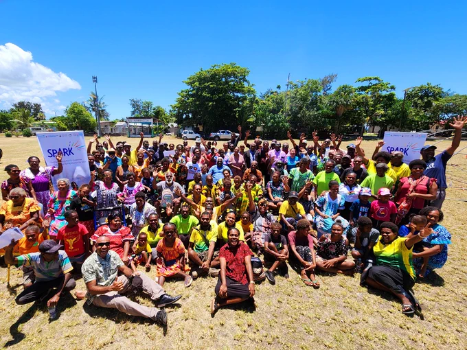A large group of community members, children, and programme participants gather outdoors under a bright blue sky during a SPARK initiative event. People wearing colourful clothing smile, wave, and pose together on a grassy field between two SPARK banners. Trees, houses, and tropical vegetation can be seen in the background, creating a vibrant community atmosphere.